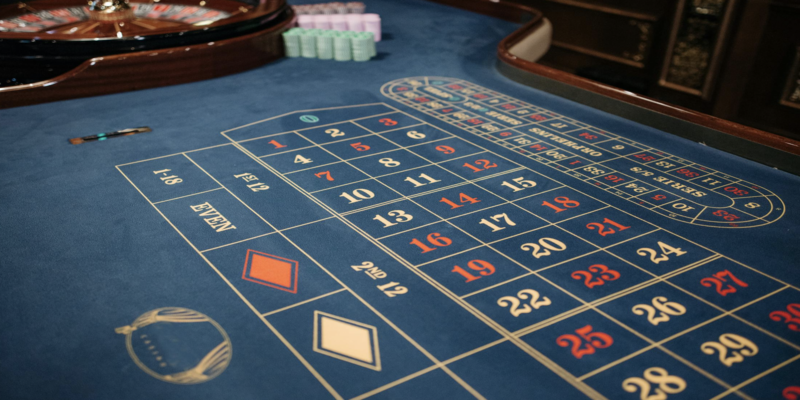 Mesa de apuestas en la que se ven los números en primer plano y una ruleta y fichas al fondo